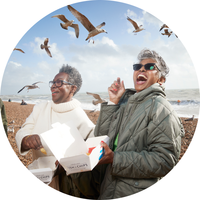 Two senior women eating fish and chips at the beach with seagulls flying overhead